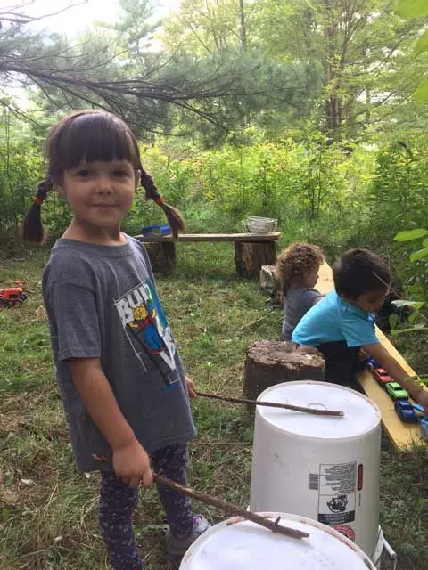 A young child with pigtails stands outdoors holding sticks near white buckets, with other children playing in the background.